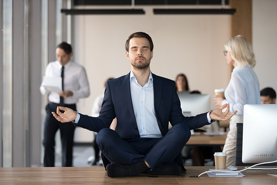 A man in a suit sits cross-legged in a yoga pose on a desk in an office