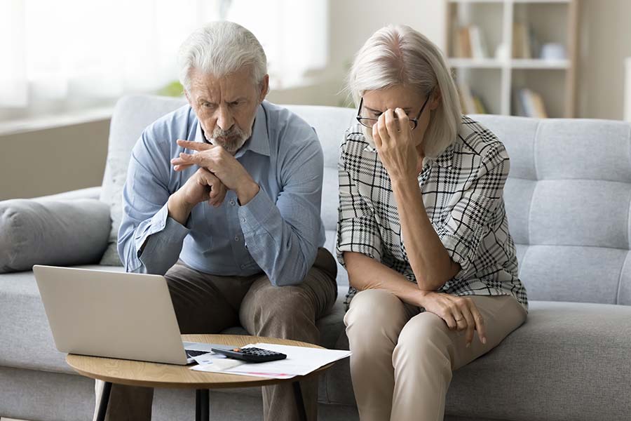 A worried-looking older couple look at a laptop screen, with a pile of papers and calculator