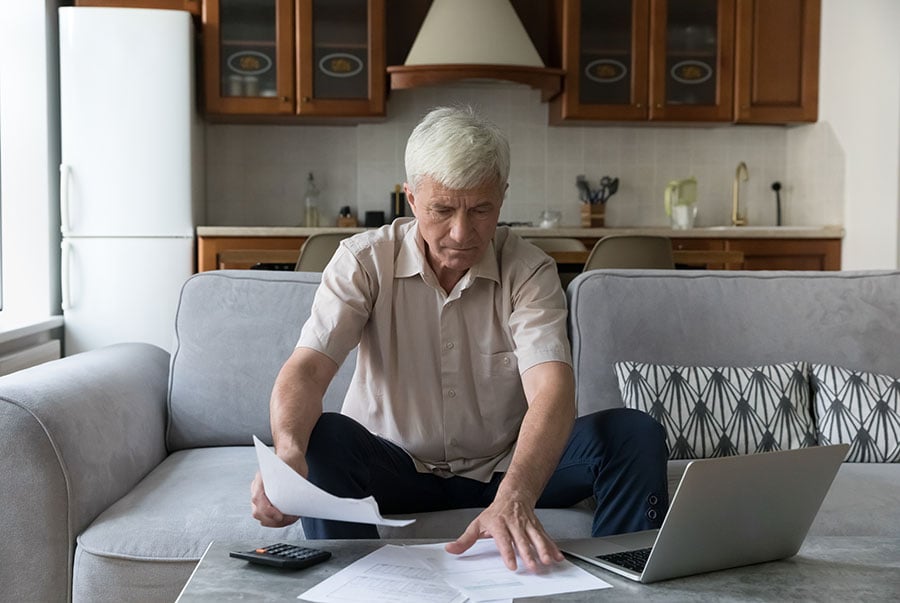 An older man sits on the sofa looking at a pile of bills