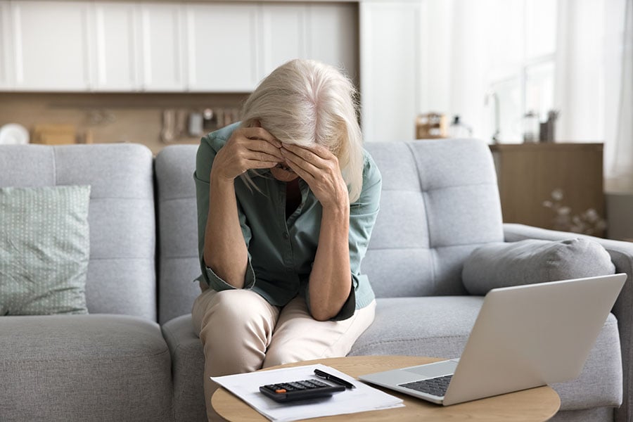 An older woman seems upset as she sits on the sofa with her head in her hands, with an open laptop in front of her