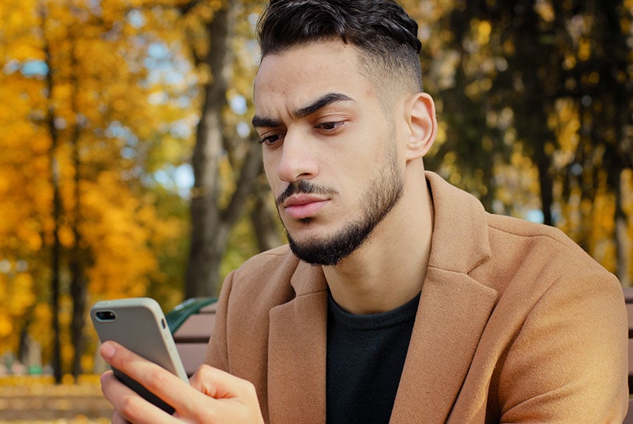 A man looks at his phone while sitting outside near trees with orange falling leaves