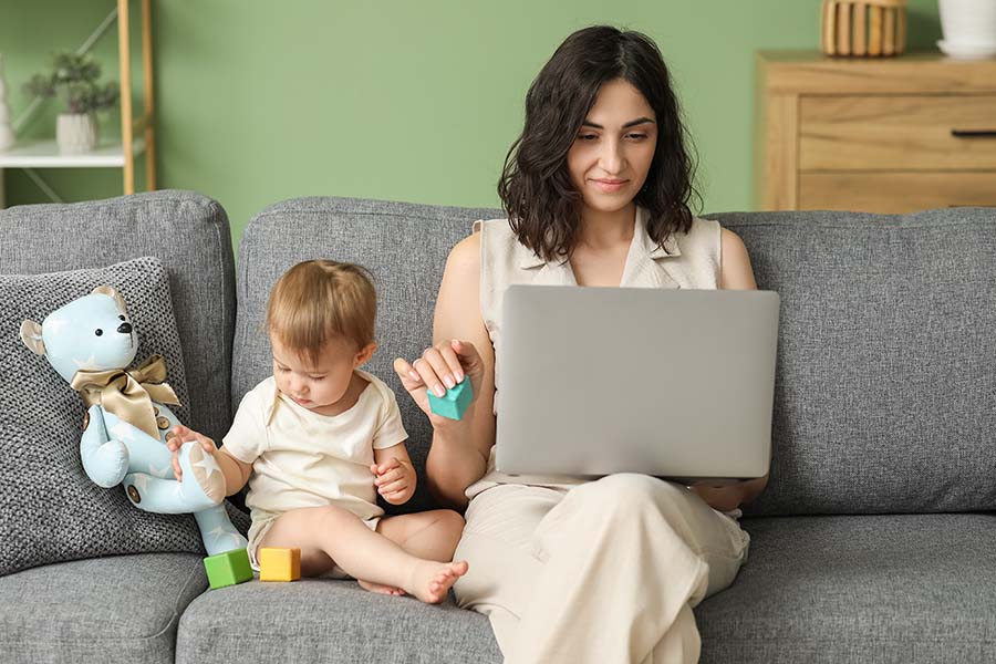A young woman sits on a sofa with an open laptop on her lap, with a baby playing next to her