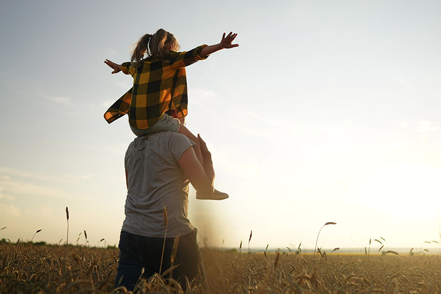 A man with a small girl on his shoulders, standing in a field at sunset