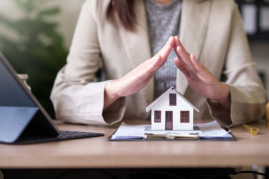 A small wooden house on a desk with a woman holding her hands protectively over the house