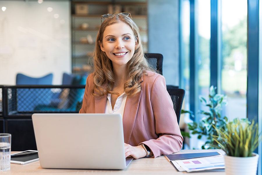 A smiling young woman sits at a desk with an open laptop