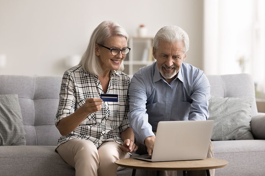 An older couple smiling as they use a laptop, while the woman holds up a credit card