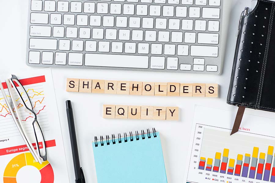 The words 'shareholders equity' on a desk surrounded by items including a keyboard, graph, notepad and reading glasses