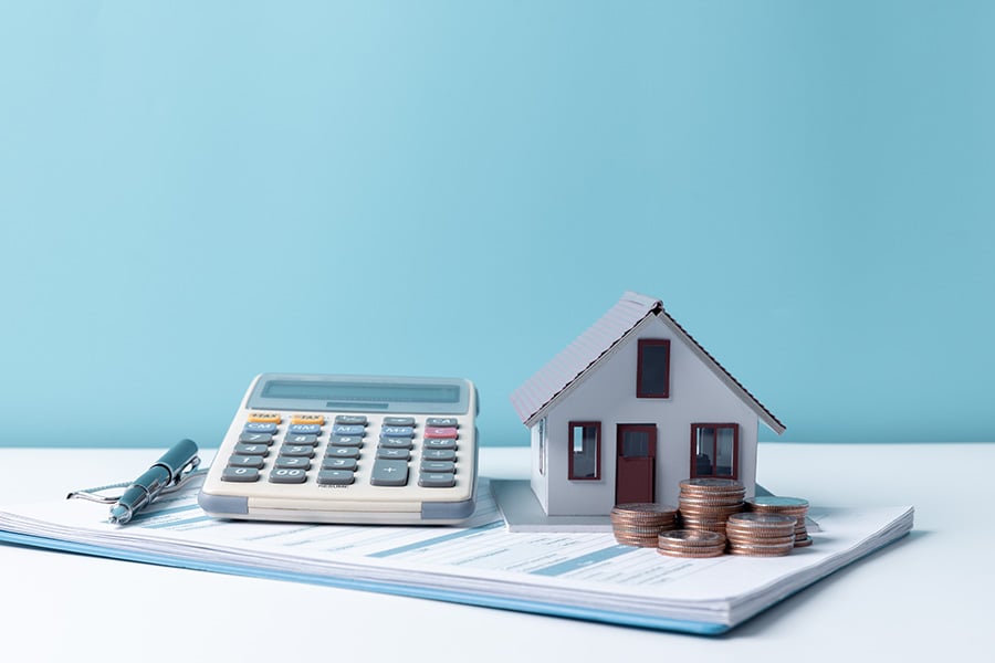 A small model house on a pile of financial statements, next to a calculator, stack of coins and a pen