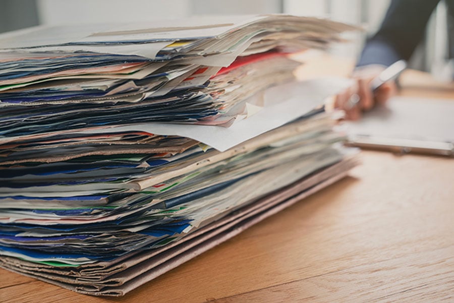A large stack of papers on a desk in the foreground, with a woman's hand holding a pan in the background