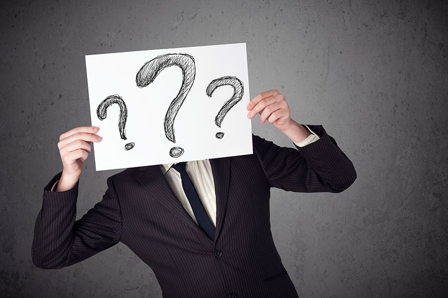A man in a black suit holds up a piece of paper with large questions marks drawn on it