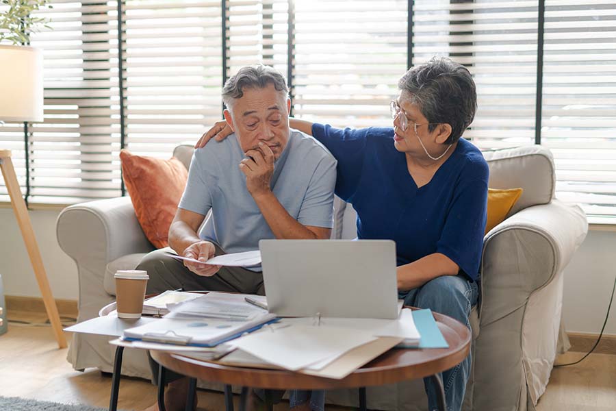 An older couple look stressed while looking at a laptop, surrounded by bills