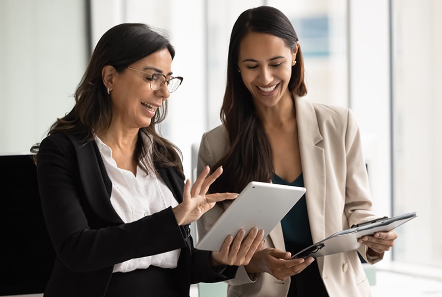 Two smiling women wearing suits in an office study a tablet computer