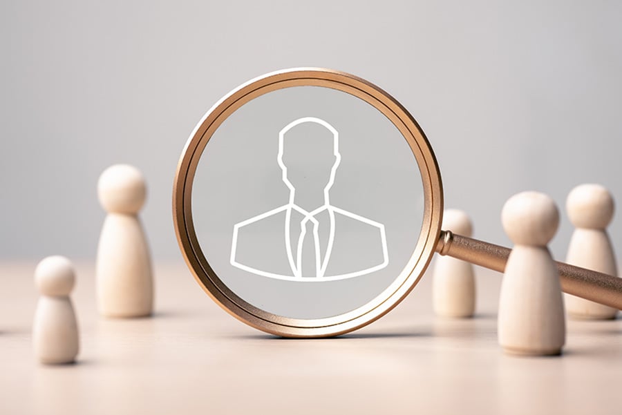 A magnifying glass over an outline of a man in a suit, surrounded by small wooden figures of people