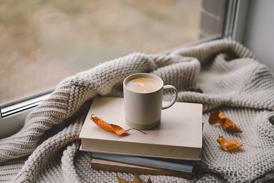 A mug of coffee on a pile of books and a blanket with autumn leaves scattered around it