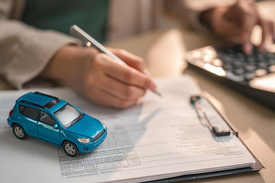 A close-up of a woman holding a pen in one hand and using a calculator with another, with a small car placed on a clipboard